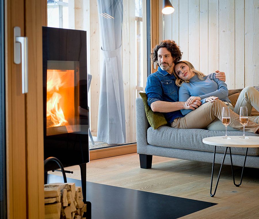 Paar entspannt auf Sofa neben modernem Kaminofen in lichtdurchflutetem Holzinterieur mit großen Fenstern