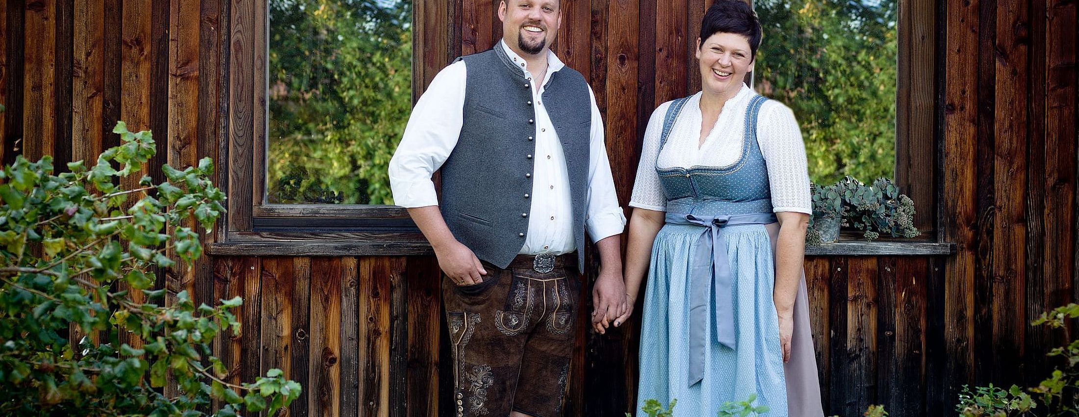 Gastgeber in bayerischer Tracht vor rustikaler Holzfassade mit Fenstern