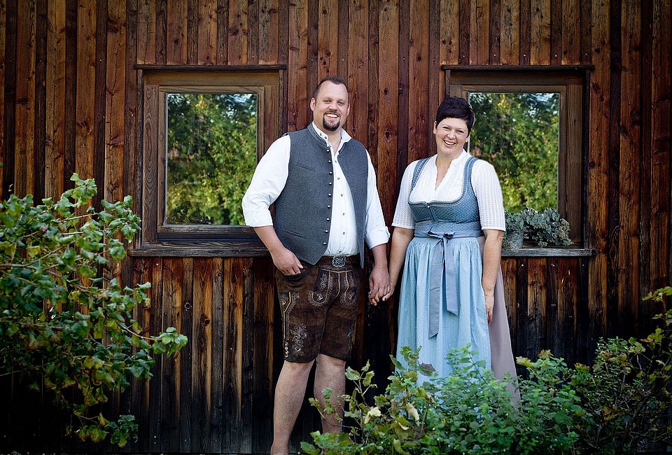 Gastgeber in bayerischer Tracht vor rustikaler Holzfassade mit Fenstern