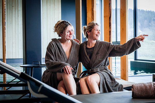 Zwei Freundinnen im Bademantel lachen auf Wellnessliegen im Ruhebereich mit Blick nach draußen