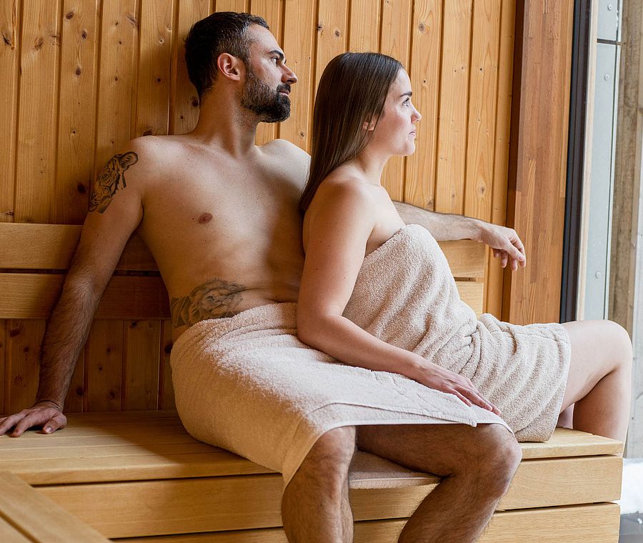 Paar sitzt in der Sauna und blickt durch das Fenster in den Schnee
